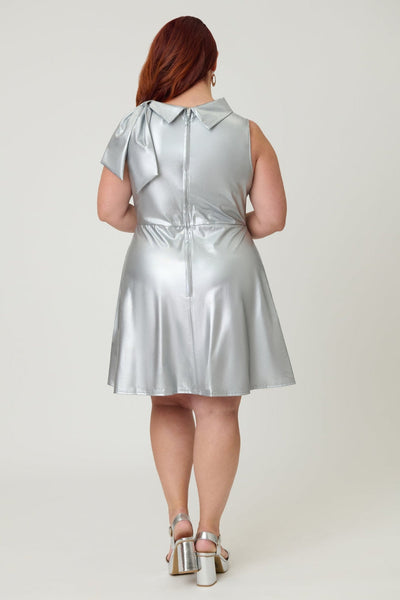 Plus size model wearing a metallic silver knit fit and flare mini dress with cowl collar neck with asymmetrical self tie detail, darting at bust, and above the knee length skirt with pockets. Shown from back