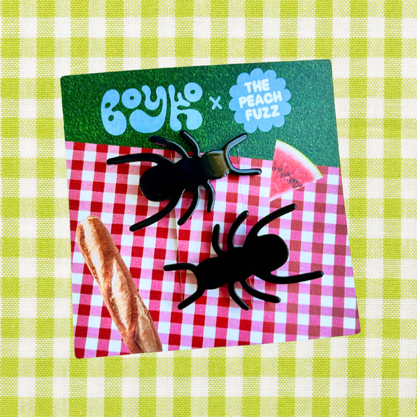 Two black acetate ant-shaped barrettes on a red and white checkered napkin with 'Boyko x The Peach Fuzz' branding on a green checkered background.
