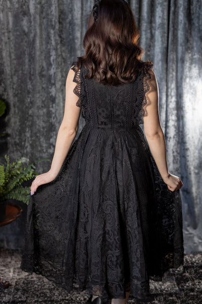 Model wearing a sleeveless midi dress with solid black shell and lace outer layer. It has a high neckline with ruffled lace collar, scalloped openwork lace at each arm opening, and a fitted bodice with three rows of geometric lace running vertically to the waist. It has a midi length lace skirt. Shown from back with skirt pulled taut
