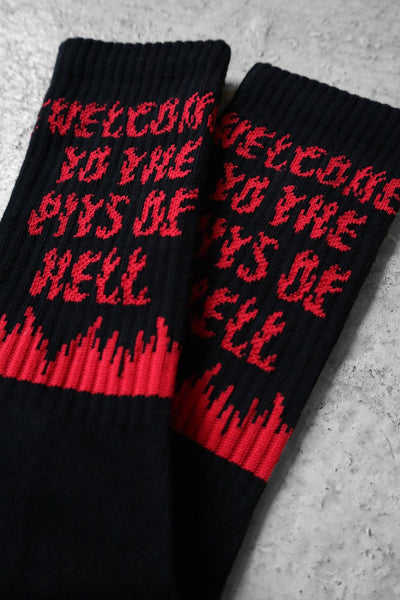 A pair of black unisex cotton crew socks with red flame accents and bold red text reading "WELCOME TO THE PITS OF HELL" and red heels and toes with "HUNGRY GHOST PRESS" writing. Shown from front in close up
