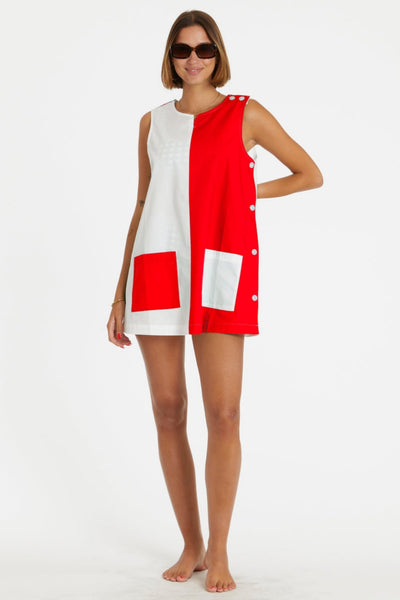 a model wearing a cotton a-line smock style mini dress in half red, half white design divided vertically down the middle of the garment. it has a round neckline with slash detail, button closure running down the left shoulder and down the side of the dress, and patch pockets in alternate colors at each hip. Shown from front