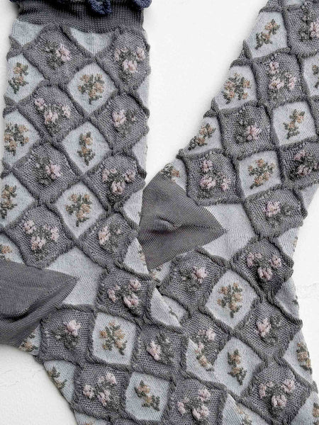 A pair of gray patterned socks with a ruffled elastic top, laid flat on a light textured background. The socks feature a diamond lattice pattern with small floral motifs in muted pink and green, and solid gray heels, toes, and cuffs. Shown flat on white background in close up