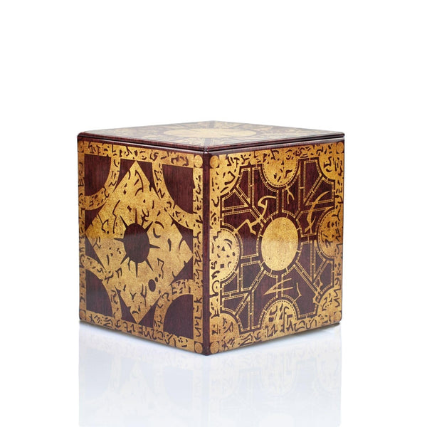 A decorative square tin box with an intricate gold and dark brown geometric and calligraphic pattern made to resemble the Lament Configuration puzzle box from Hellraiser, shown on a reflective white surface against a white background.