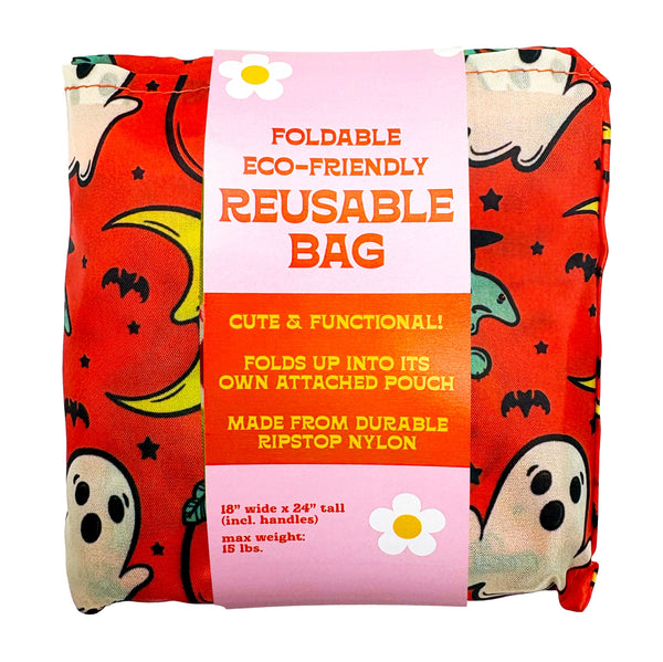 An orange reusable nylon tote bag with an all-over pattern of cartoonish retro Halloween witches, ghosts, crescent moons, and jack-o'-lanterns shown wrapped in a pink paper band label that reads 'FOLDABLE ECO-FRIENDLY REUSABLE BAG'.