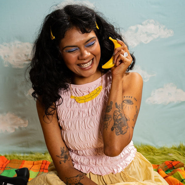 model wearing marbled acetate necklace with large horizontal charm in the shape of a banana with black spots and scattered rhinestone adornment on a gold paperclip style chain. 