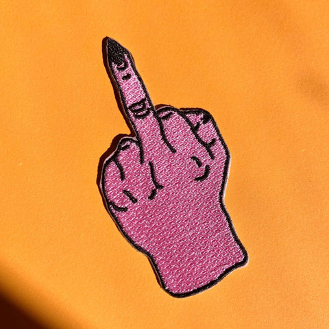An embroidered patch of a bright pink hand giving the middle finger with pointy black fingernails and black detailing. Shown flat on an orange background 