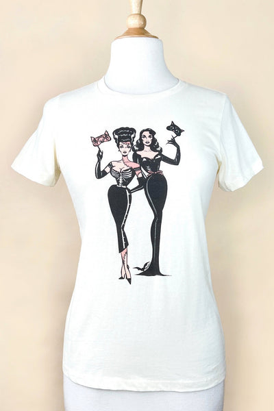 ivory white t-shirt with printed illustration of two pinup-style women holding masquerade masks, one dressed as the Bride of Frankenstein and the other dressed as a black cat. Shown from front on dress form