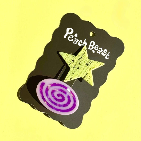 A set of two acetate barrettes, one in a chartreuse glittery star shape and a purple spiral with lavender background oval shape, set against a solid pale yellow background and shown on black backing card