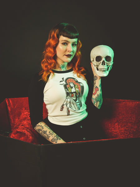 off-white crop length black raglan shoulder top with round neck, three quarter sleeves, and printed illustration of pinup style woman with stitched legs wearing gown and sitting in coffin-style large chair. Printed in red, black, blue, and yellow. Shown worn by model sitting in coffin holding skull