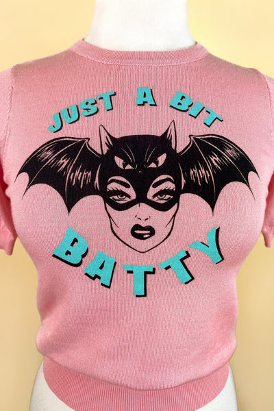 pink crew neck short sleeved sweater with retro woman wearing a bat mask and “Just a Bit Batty” written in bright blue. Shown on dress form in closeup