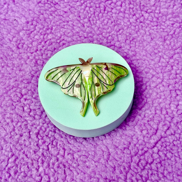 Green and brown iridescent acetate French style barrette in the shape of a Luna Moth. Shown on a fuzzy purple background