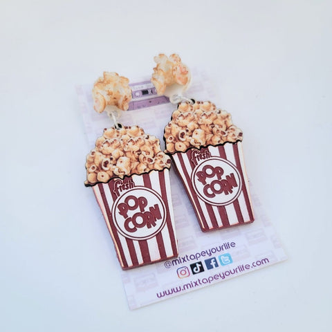 laser-cut acrylic drop earrings in the shape of red and white striped boxes of popcorn connected to a 3D resin popcorn charm. Shown on backing card