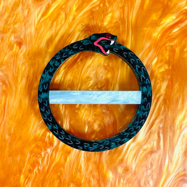 laser-cut black and green marbled layered acrylic scarf slide with hand painted scale and fang details of an ouroboros. Shown on marbled orange background