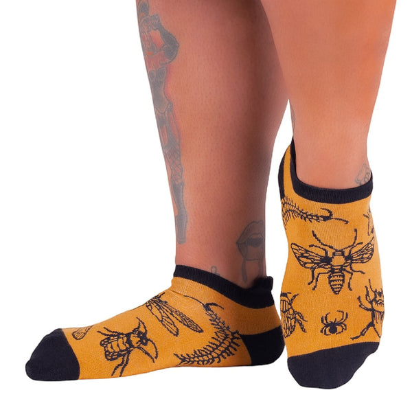 mustard unisex ankle socks with black toes,heels, and cuffs with an all over pattern of bug illustrations with black outlines including scorpions, dragonflies, centipedes, moths, and spiders. Shown worn by a model from the front