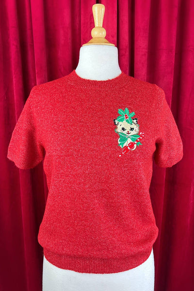 Red sweater with red lurex threads with an embroidered white cat with holly, a green bow, and red mittens holding a candy cane design on a mannequin against a red curtain
