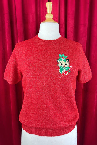 Red sweater with red lurex threads with an embroidered white cat with holly, a green bow, and red mittens holding a candy cane design on a mannequin against a red curtain