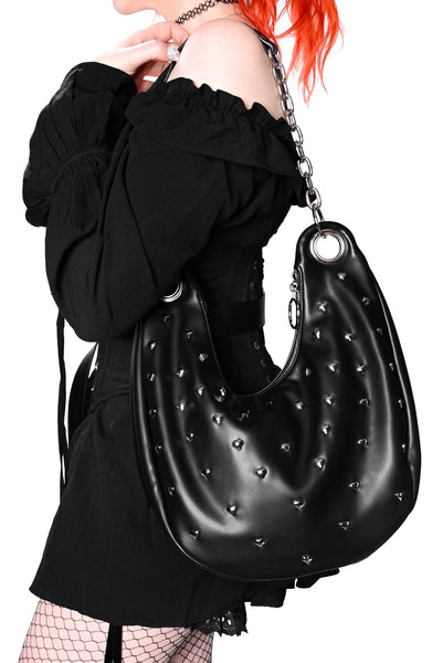 Black heart shaped studded handbag with silver chain strap, oversized silver grommets, and o-ring top zip closure. Shown held on model's shoulder from side