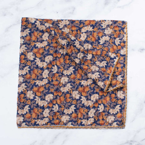 square seersucker cotton scarf with navy blue background and allover floral pattern of leaves, flowers, and berries in cream, orange, and brown. Embroidered scalloped border in mustard yellow