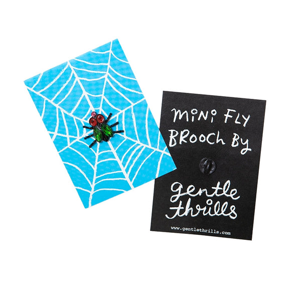 small shiny black enameled house fly-shaped brooch with enameled red eyes, almond-shaped bright green jewels for wings, an a small light pink jewel for a nose. Shown from front and back on backing card