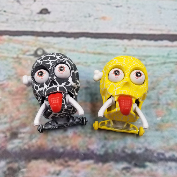 Two wind-up toy skull figures, one black and white with red tongue, the other yellow with red tongue, both with bloodshot eyes, on a textured surface.