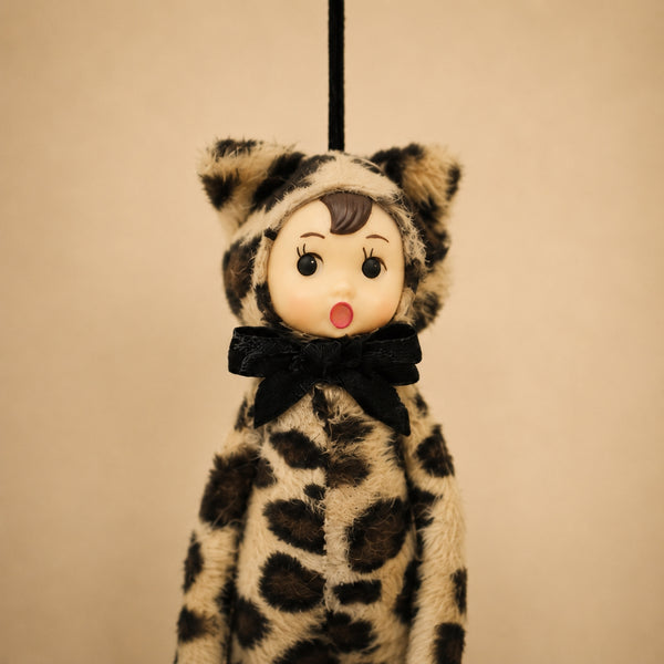 Poseable ornament with wire limbs and Velcro hands in the shape of a pixie with rosy cheeked, green eyed resin face with molded tuft of brown hair. She is wearing a faux fur leopard costume with ears and tail. Black satin ribbon for hanging is at top of head. Shown in close up on beige background