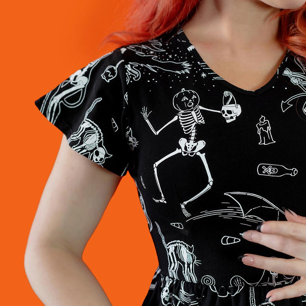 Model wearing a black cotton jersey knit dress with white skeleton, owl, pumpkin, witch and bat print. Dress has a shallow v-neckline, short flutter sleeves, a knee length double tiered skirt with ruffle hem and pockets. Shown from front against an orange background in close up of sleeve and print detail 