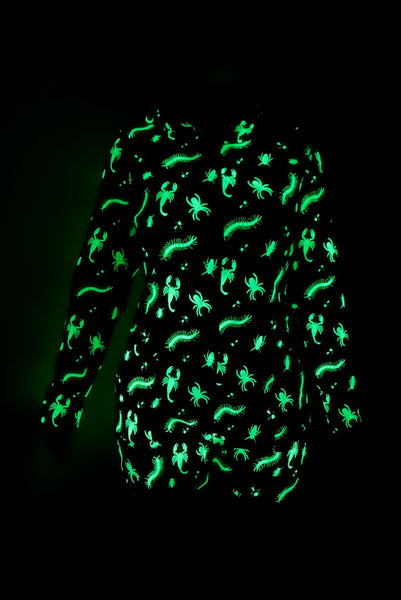 model wearing a black long sleeve hooded onesie with neon green insect pattern, front zip closure, and mid-length shorts. Shown from front in dark to display intense glow of print