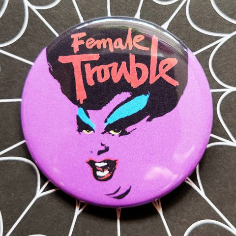 1.25" round pinback button with neon purple background and black & blue illustration of Divine with "Female Trouble" written in red