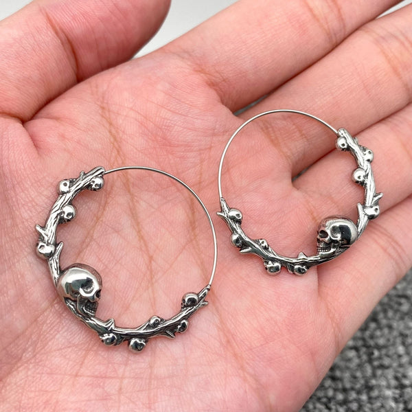 stainless steel infinity closure hoop earrings with skull and branch detail at the bottom of each hoop with tiny skulls growing out of each branch. Shown held in palm of hand