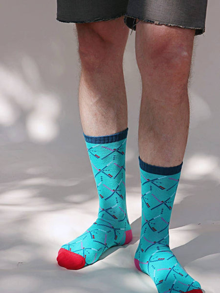 Pair of unisex crew socks with a knit-in pattern of the geometric carpet design of the Portland International Airport. With a teal background, navy blue cuffs, pink heels, and red toes. Shown worn by a model on a grey background 