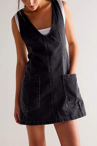 Black denim jumper style dress with v-neckline, vertical seaming, and large patch pockets worn by a person on a plain background