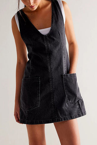 Black denim jumper style dress with v-neckline, vertical seaming, and large patch pockets worn by a person on a plain background