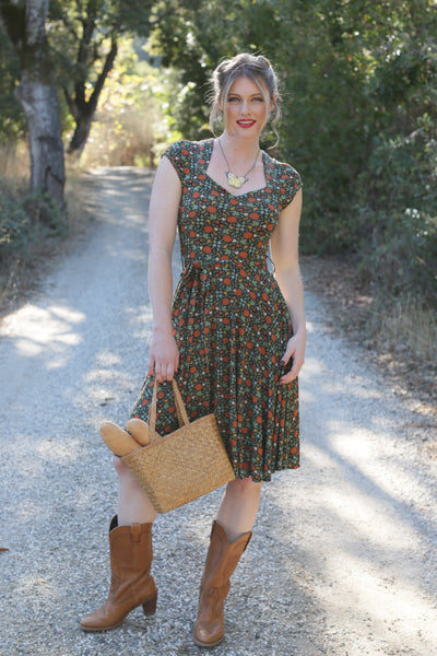 model in an outdoor setting wearing a black background dense stylized autumnal multi orange, green, and cream floral pattern fit and flare dress featuring a Queen Anne neckline, cap sleeves, swingy just-below-the-knee length skirt