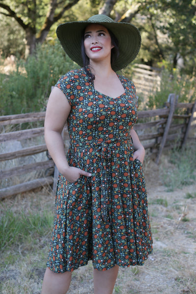 model in an outdoor setting wearing a black background dense stylized autumnal multi orange, green, and cream floral pattern fit and flare dress featuring a Queen Anne neckline, cap sleeves, swingy just-below-the-knee length skirt