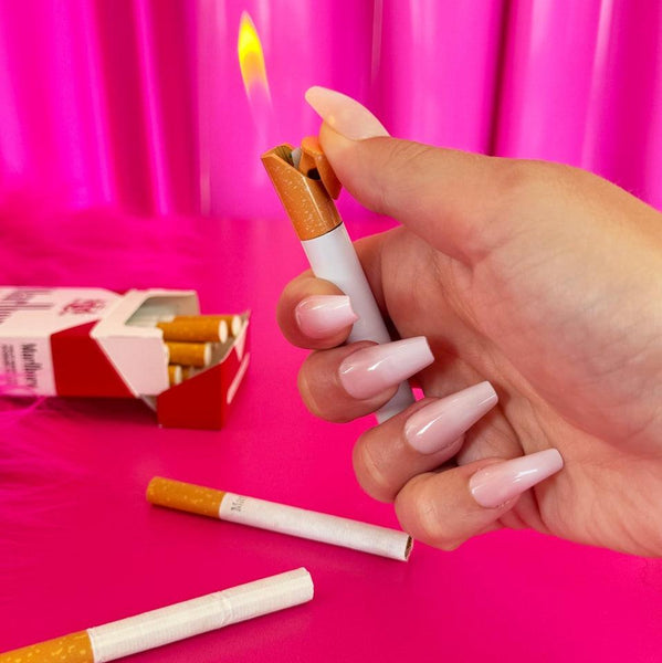 yellow and brown lighter in the shape of a cigarette. Held in front of a pink background with flame alight