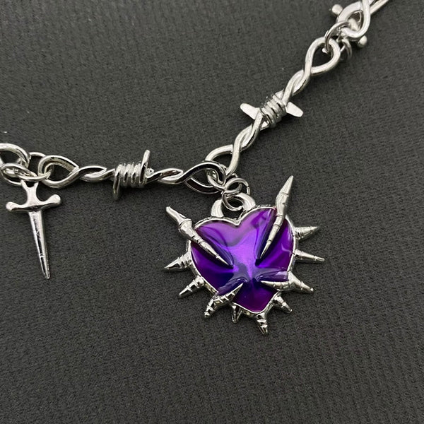 Silver necklace with barbed wire style chain, dangling tiny dagger pendants, and purple enameled spiked heart pendant shown in close up on a dark background