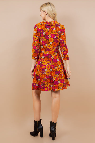model wearing a knit long sleeved dress in a brown background pink, purple, orange, and white retro daisy print . it has a cowl style neck, pockets, and knee length skirt. Shown from the back