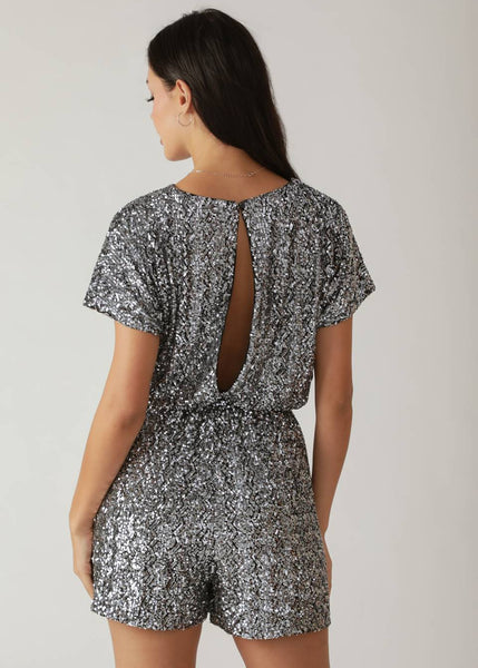model wearing a short sleeved romper in silver sequin fabric with zigzag stripe pattern, round neckline, gathered elasticized waist, and deep keyhole detail at back. Shown from back
