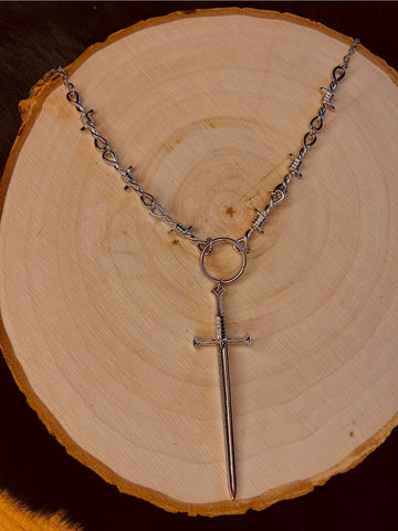 24” silver metal barbed wire necklace with a sword charm connected by a metal o ring. Shown flat on a tree stump