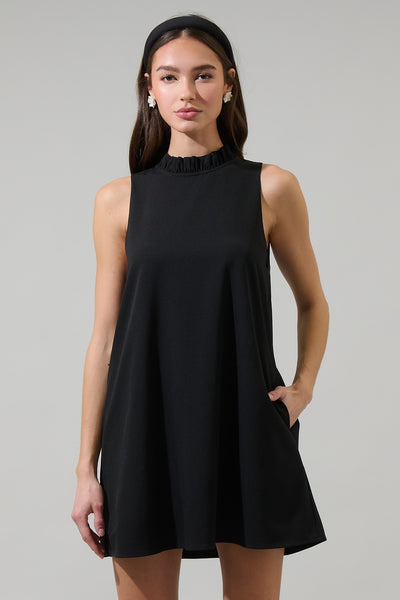 model wearing a black woven sleeveless trapeze dress with high ruffled neckline, pockets, skirt ending above the knee, and self tie at nape of neck. Shown from front