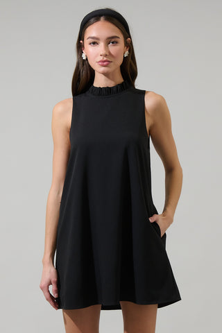 model wearing a black woven sleeveless trapeze dress with high ruffled neckline, pockets, skirt ending above the knee, and self tie at nape of neck. Shown from front