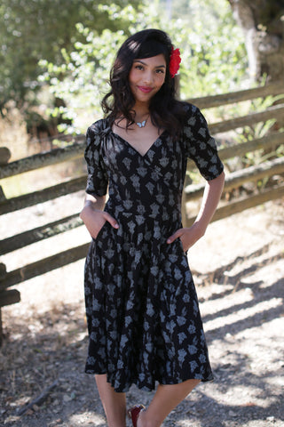 black stretch cotton fit and flare dress in an allover printed ornate cuckoo clock pattern in cream featuring a surplice bodice with Queen Anne neckline, puffed shoulder short sleeves, and full knee length skirt. shown worn by a model in an outdoor setting