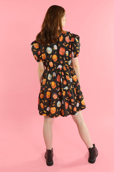 model wearing a babydoll style dress with a black background photorealistic collage pattern of doll heads and arms. It has a ruffled Peter Pan style collar, puffed short sleeves, and a gathered double tiered above the knee skirt. Shown from the back
