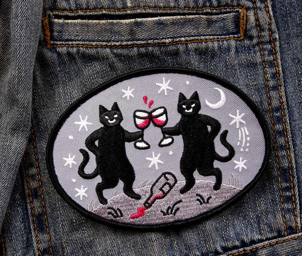 Oval twill embroidered patch of two black cats holding glasses of red wine with a spilled bottle at their feet surrounded by white moons and stars on a grey background 