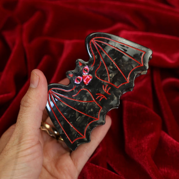 black marbled acetate claw style hair clip in the shape of a bat with outstretched wings and red and pink mouth with red line detail. Shown held flat on red velvet background for scale