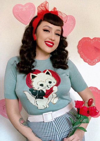 A model wearing a powder blue short sleeved sweater with a printed illustration of a white kitten with a red valentine heart around its head and a black bow around its neck.