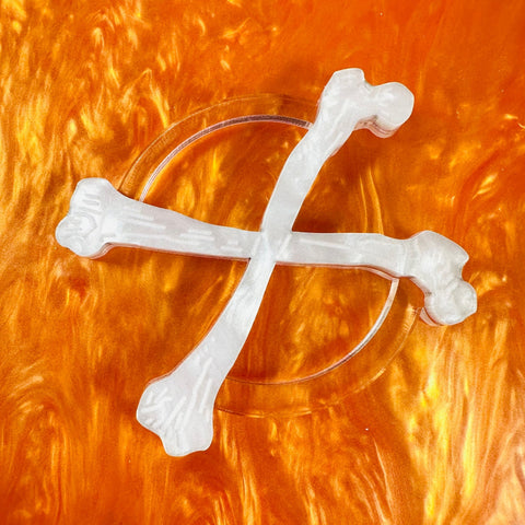 white pearly laser cut acrylic scarf slide in the shape of a pair of crossed bones with etched detail and clear backing shown on pearly orange background