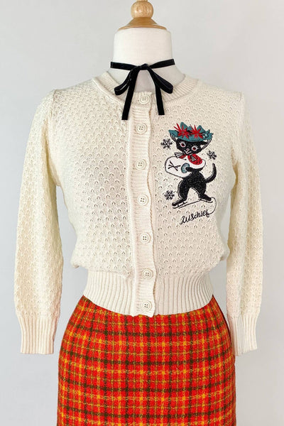 warm white cotton pointelle cardigan with large embroidery on left chest of black cat wearing ice skates, green hat with poinsettias, and white muff alongside trio of snowflakes and "Mischief" written in script below. Shown on dress form from front