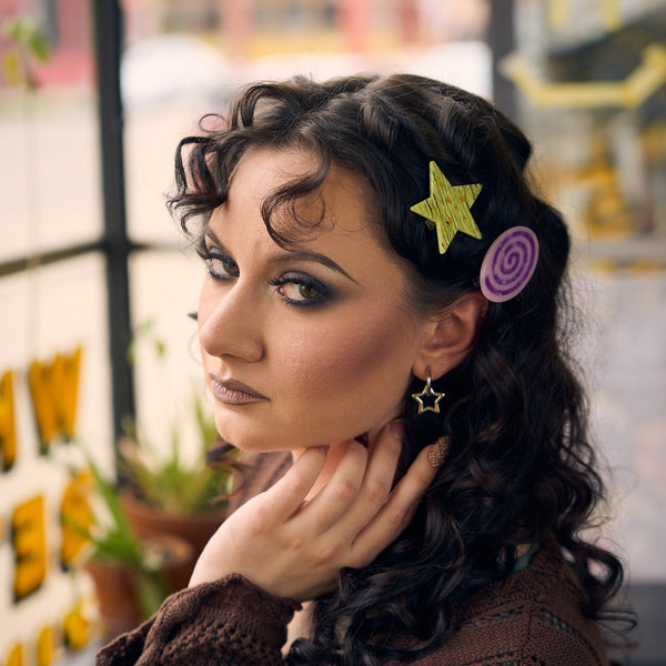 A set of two acetate barrettes, one in a chartreuse glittery star shape and a purple spiral with lavender background oval shape, shown worn by model with curly hair