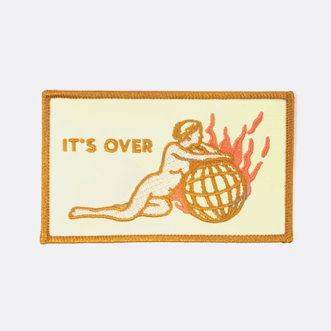 rectangular pale yellow patch with burnt orange border and nude feminine figure reclining against a burning planet with message "IT'S OVER"